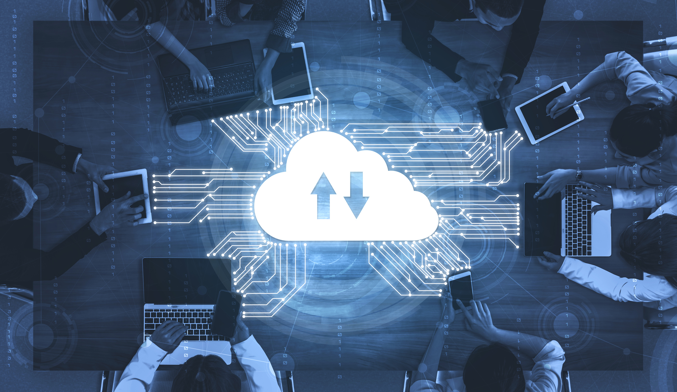 An overhead view of a group of professionals working together with laptops and tablets on a wooden table, surrounded by digital graphics representing cloud services, including a cloud icon with upward and downward arrows, symbolizing data transfer.